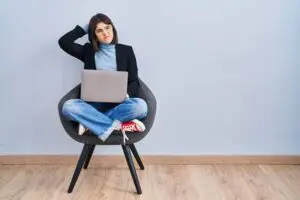 How smart is using AI in recruitment? 18 Young hispanic woman sitting on chair using computer laptop confuse and wondering about question. uncertain with doubt, thinking with hand on head. pensive concept.