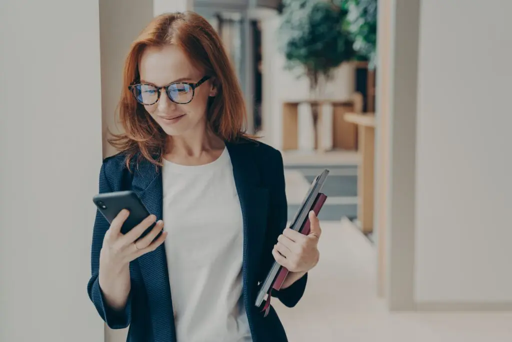 Elegant smiling woman office worker in eyeglasses using modern mobile phone, sending message or chatting with client online, standing in coworking space with laptop and note book in hand