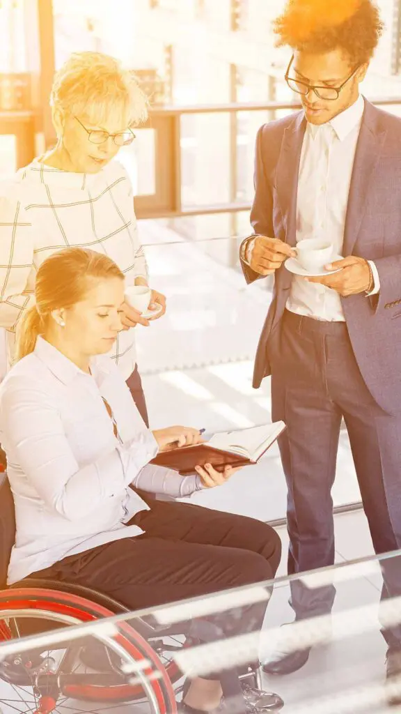A diverse group of people talking, one is in a wheelchair holding a book and a male and female are looking down to the book where she is pointing.