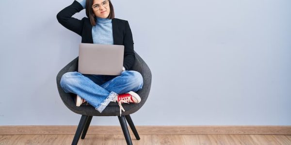 Young hispanic woman sitting on chair using computer laptop confuse and wondering about question. uncertain with doubt, thinking with hand on head. pensive concept.